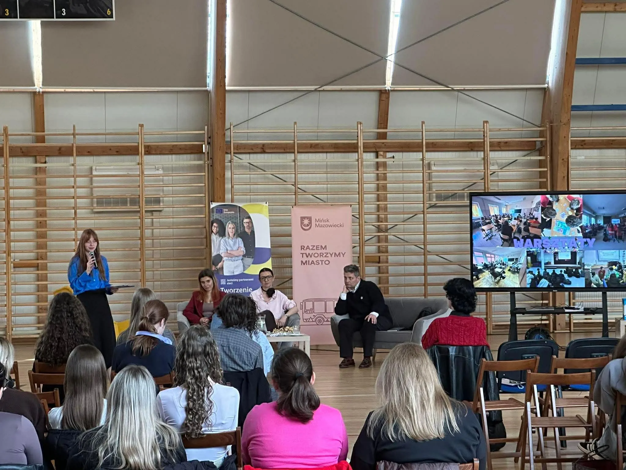 Sala gimnastyczna z drewnianymi drabinkami na ścianach. Publiczność siedzi w rzędach skierowana na scenę. Po lewej kobieta w niebieskiej bluzce przemawia do mikrofonu. Na środku trzy osoby siedzą na kanapie obok stojących banerów. Po prawej stoi duży ekran z wieloma miniaturami zdjęć.