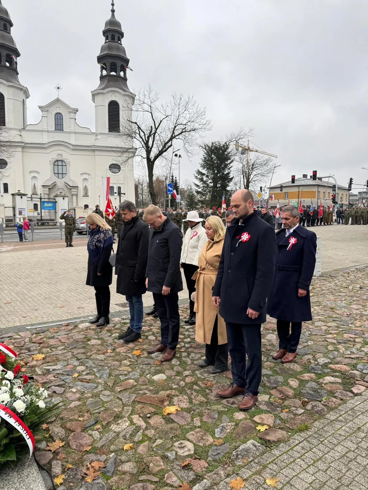 Siedem osób stoi na kamiennej nawierzchni głową skierowaną w dół, oddając hołd. W tle widać białą budowlę z dwiema wieżami oraz grupę wojskowych i flagi. Niebo zachmurzone, dzień.