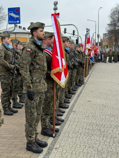 Rząd wojskowych w umundurowaniu zimowym stoi w szeregu na chodniku przy ulicy, kilku żołnierzy trzyma czerwone i białe sztandary z orłem państwowym, w tle sygnalizacja świetlna i przejście dla pieszych, dzień, atmosfera formalna.