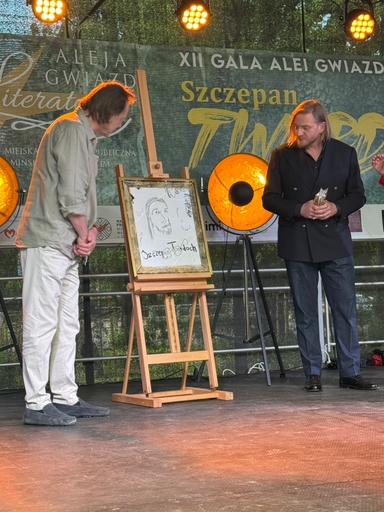Dwóch mężczyzn stoi na scenie przy sztaludze z oprawionym rysunkiem. Za nimi świecą pomarańczowe reflektory, a na tle wisi zielony baner z dużymi napisami. Wydarzenie odbywa się na zewnątrz w ciągu dnia.