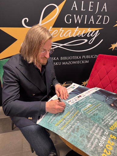Mężczyzna z włosami do ramion i okularami siedzi przy stole wewnątrz i podpisuje plakat. Za nim czarne tło z dużą żółtą gwiazdą i białym napisem. Po prawej czerwone pikowane krzesło.