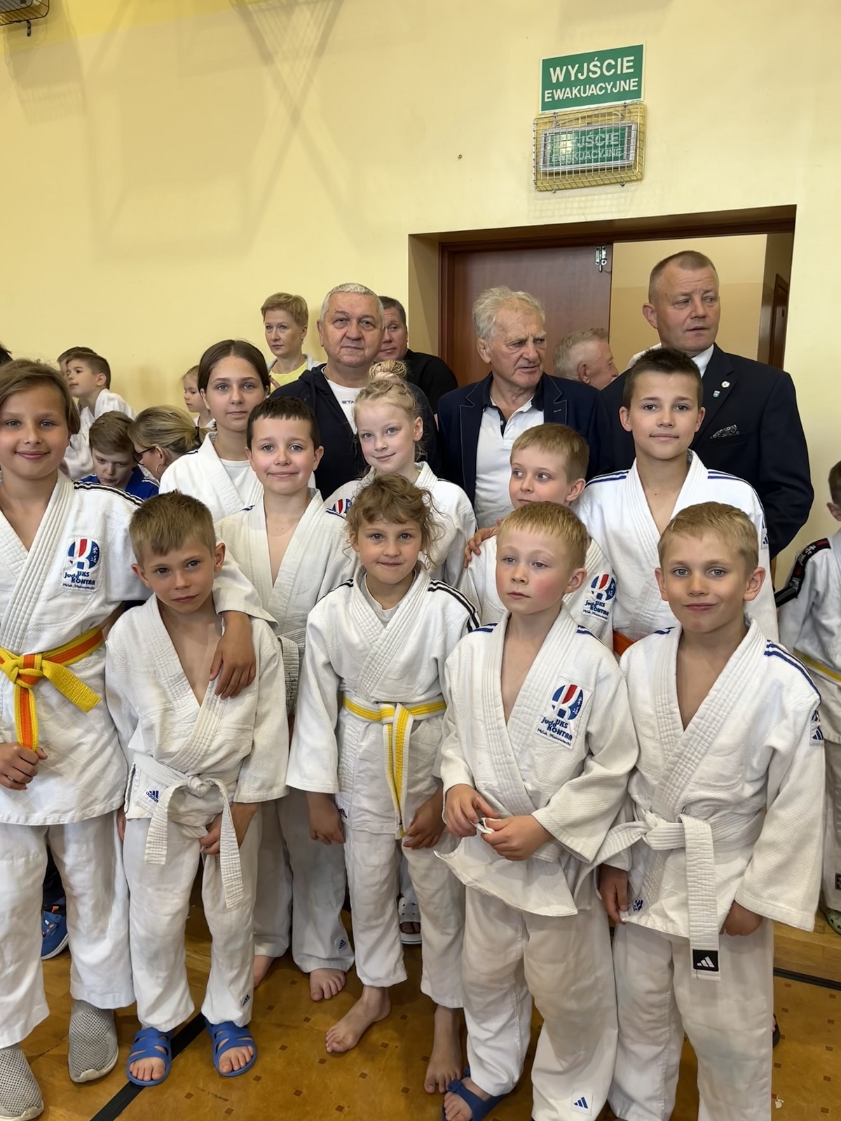 Około dziesięcioro dzieci w białych judogach stoi blisko siebie na drewnianej podłodze hali sportowej. Kilku dorosłych mężczyzn stoi za dziećmi przy otwartym przejściu w żółtej ścianie. Na ścianie nad przejściem widoczny zielony znak awaryjny. Każde dziecko patrzy w stronę aparatu.