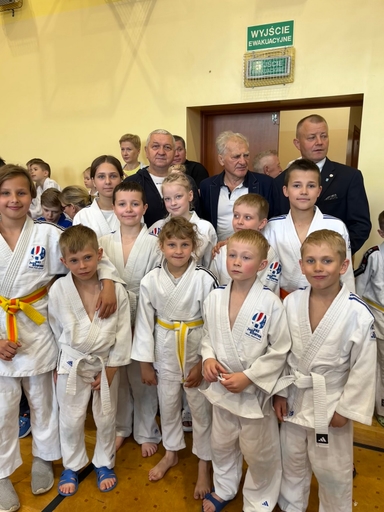 Około dziesięcioro dzieci w białych judogach stoi blisko siebie na drewnianej podłodze hali sportowej. Kilku dorosłych mężczyzn stoi za dziećmi przy otwartym przejściu w żółtej ścianie. Na ścianie nad przejściem widoczny zielony znak awaryjny. Każde dziecko patrzy w stronę aparatu.
