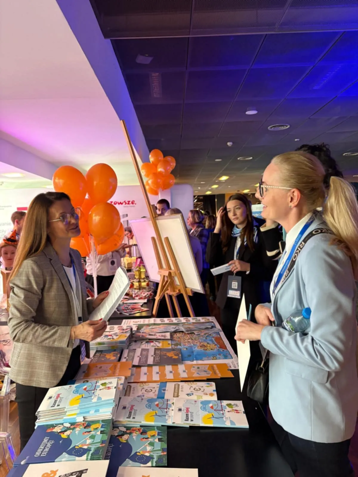 Dwie kobiety rozmawiają przy stoisku z ulotkami i broszurami. W tle stoi kilka osób, na stoisku znajdują się pomarańczowe balony. Wnętrze z ciemnym sufitem, oświetlenie mieszane, strona lewa jasna, prawa w niebieskim świetle.