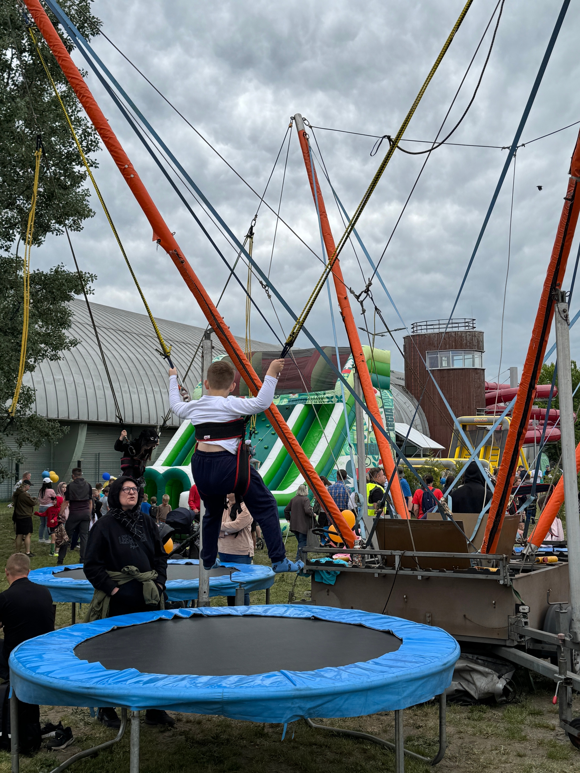 Dziecko w uprzęży stoi na trampolinie podpięte do lin i pomarańczowych masztów. W tle duża zielono-biała dmuchana zjeżdżalnia, grupa ludzi i budynek z okrągłą wieżą. Niebo jest pochmurne, sceneria odbywa się na zewnątrz w dzień.