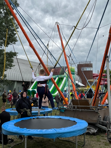 Dziecko w uprzęży stoi na trampolinie podpięte do lin i pomarańczowych masztów. W tle duża zielono-biała dmuchana zjeżdżalnia, grupa ludzi i budynek z okrągłą wieżą. Niebo jest pochmurne, sceneria odbywa się na zewnątrz w dzień.