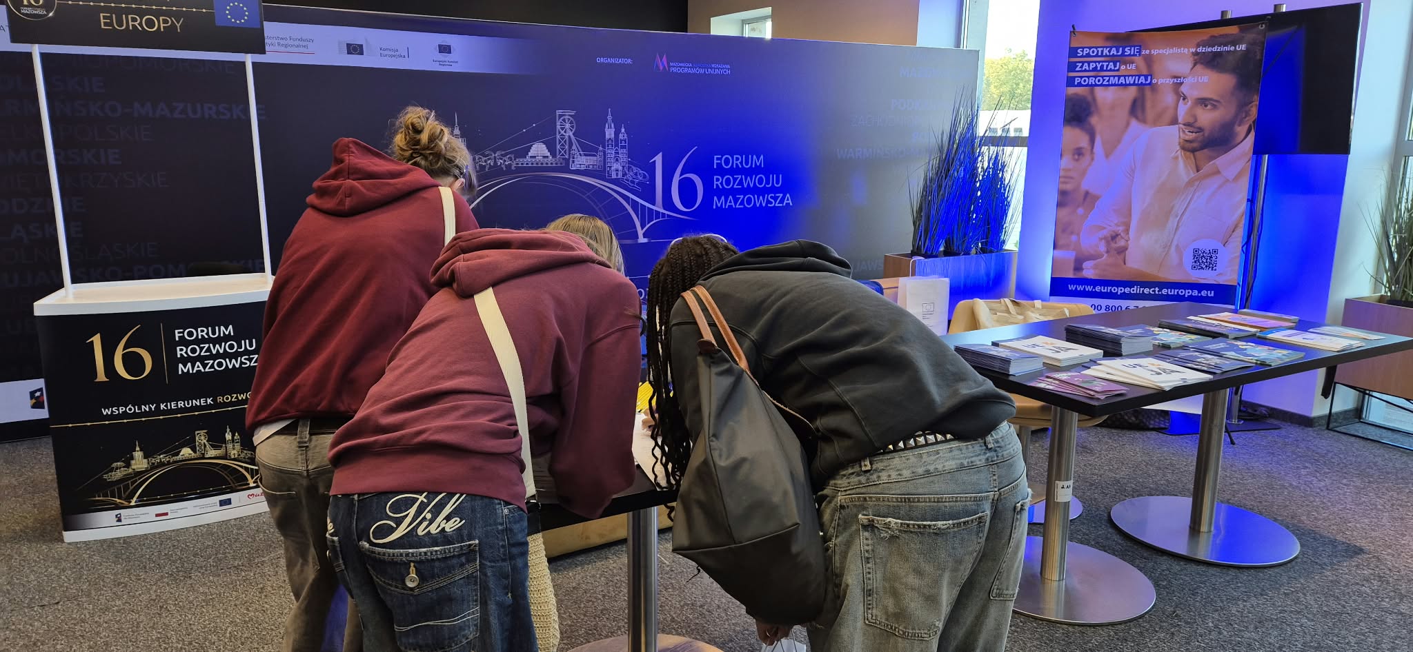 Trzy osoby pochylone nad stołem informacyjnym w hali konferencyjnej. Dwie mają bordowe bluzy, jedna czarną kurtkę. Na stole materiały informacyjne i ulotki. W tle duże niebieskie ścianki z grafiką miasta i jasnym oświetleniem. Pomieszczenie jest wnętrzem, widoczne stojaki i rośliny.