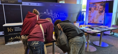 Trzy osoby pochylone nad stołem informacyjnym w hali konferencyjnej. Dwie mają bordowe bluzy, jedna czarną kurtkę. Na stole materiały informacyjne i ulotki. W tle duże niebieskie ścianki z grafiką miasta i jasnym oświetleniem. Pomieszczenie jest wnętrzem, widoczne stojaki i rośliny.