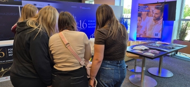 Cztery osoby stoją tyłem do kamery przy stoisku wystawowym w pomieszczeniu. Tło ma duży niebieski baner z napisem „16 Forum Rozwoju Mazowsza”. Po prawej stoi stolik z ulotkami i stojak reklamowy. Widać okno z zielonymi roślinami i dzienne światło.