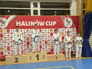 Pięciu chłopców w judogach stoi na podium i obok niego w hali sportowej z niebieską podłogą i czerwoną ścianką z napisem HALINÓW CUP oraz wieloma logotypami. Trzech chłopców stoi na podium oznaczonym numerami 1, 2 i 3, trzej trzymają dyplomy i medale. Dwaj chłopcy stoją po prawej stronie podium, również trzymają dyplomy.