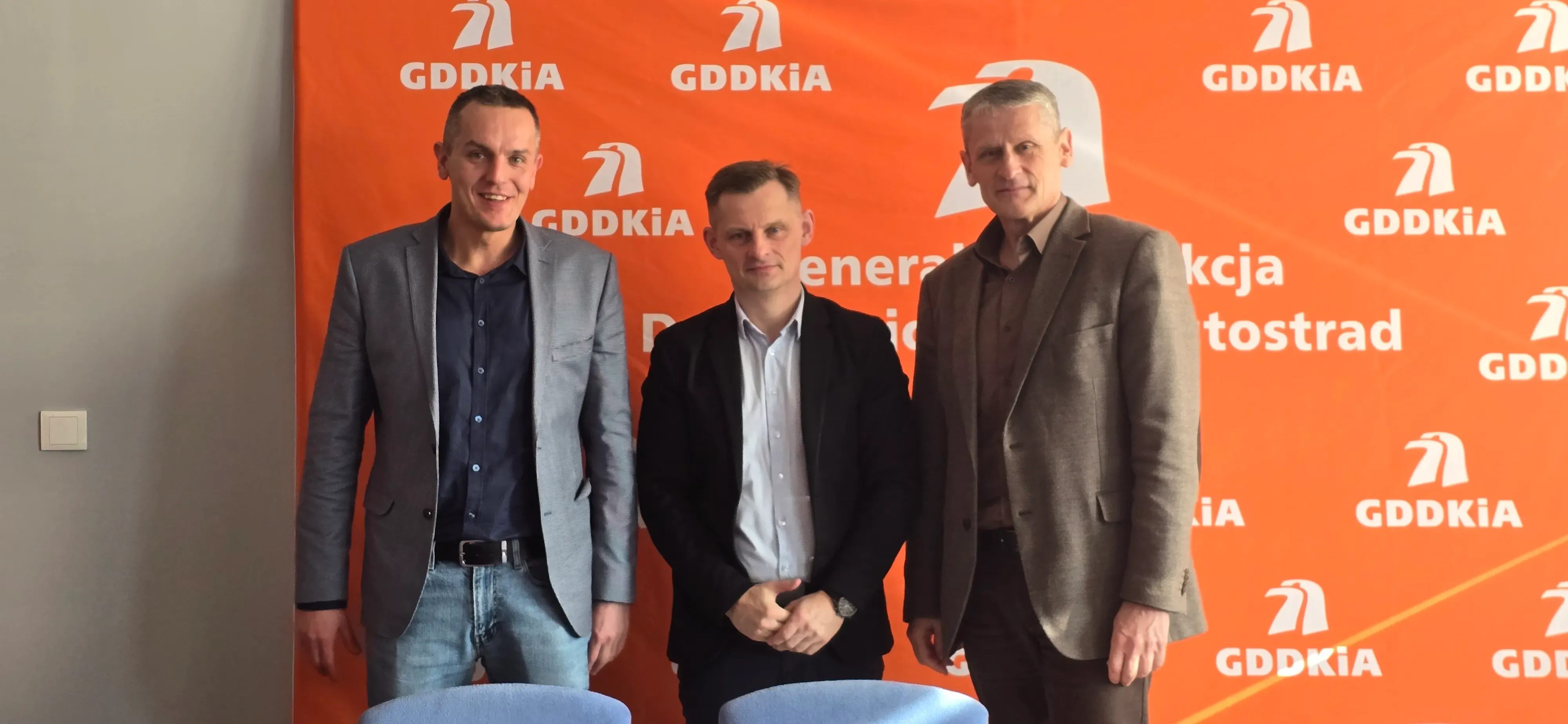 Wnętrze budynku. Trzech mężczyzn stoi przed pomarańczowym tłem z powtarzającymi się białymi logotypami. Mężczyzna po lewej ma szarą marynarkę i dżinsy. Stojący w środku ma ciemną marynarkę i jasną koszulę. Mężczyzna z praerj ma brązowy garnitur. Pozują do zdjęcia.