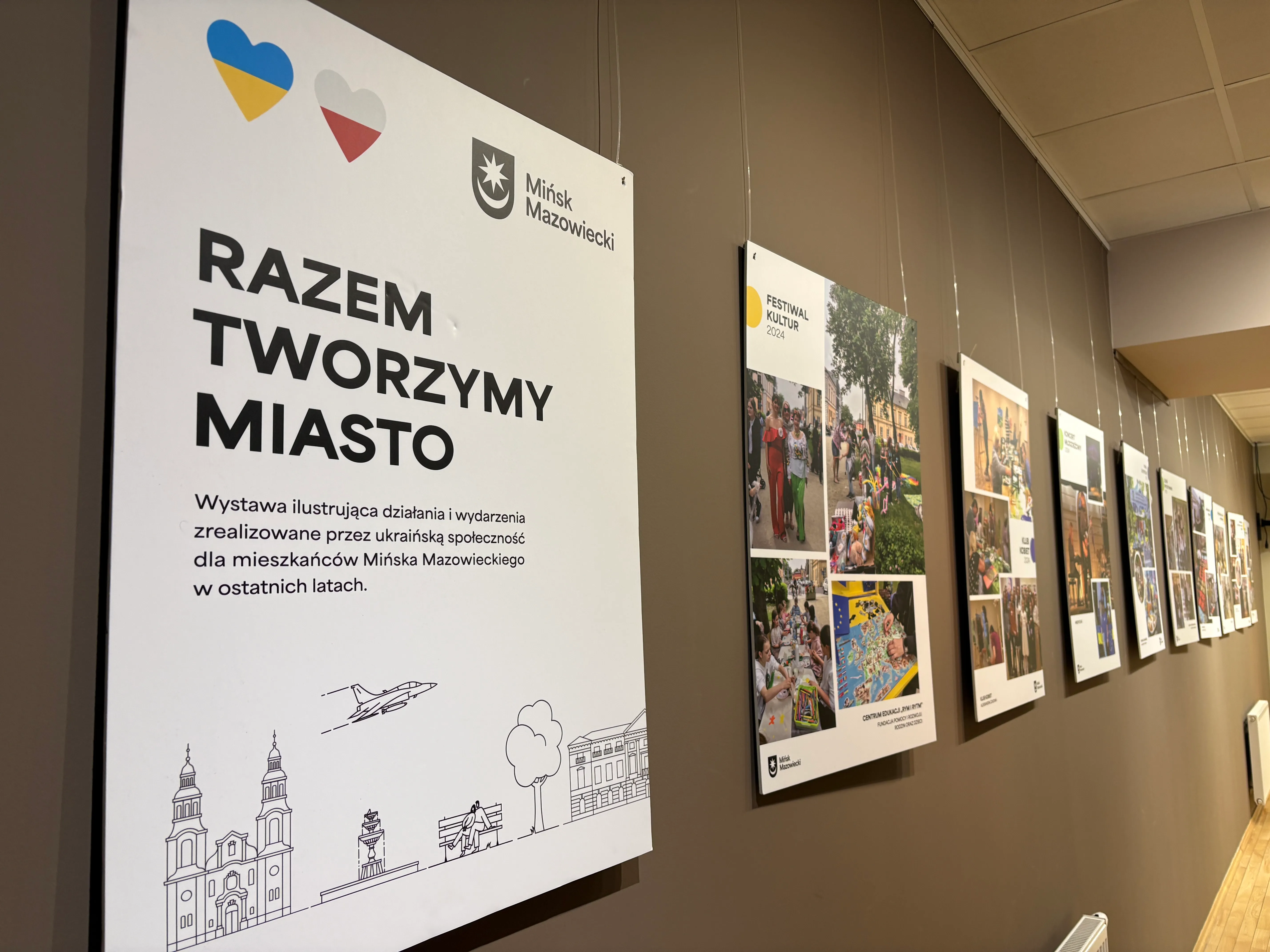 Duży biały panel informacyjny z dwoma kolorowymi sercami w lewym górnym rogu zamocowany na brązowej ścianie. Po prawej stronie kilka wiszących plansz z kolażami fotografii ustawionych w rzędzie. Wnętrze galerii z podwieszanym sufitem i grzejnikiem przy podłodze.