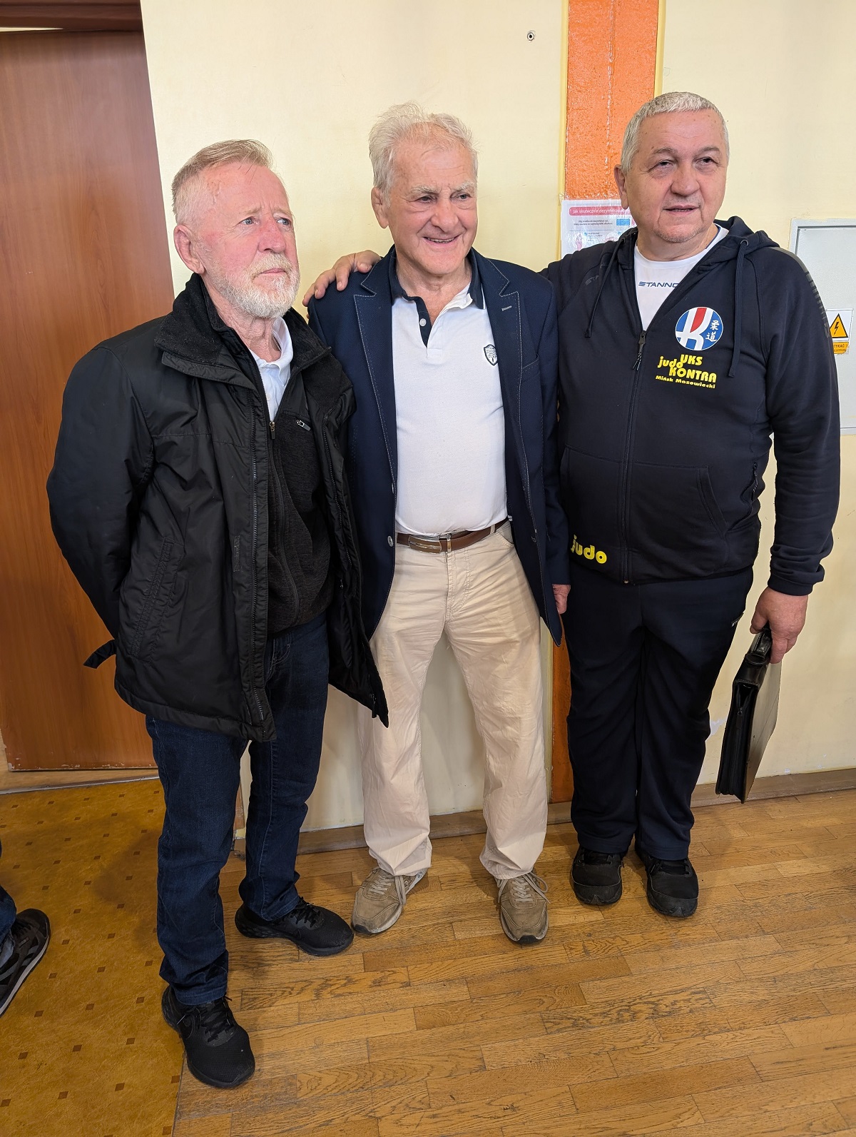 Trzech starszych mężczyzn stoi wewnątrz przy jasnobeżowej ścianie z pomarańczowym pionowym pasem. Środkowy mężczyzna uśmiecha się i ma ręce na ramionach pozostałych. Lewy mężczyzna ma siwą brodę i czarną kurtkę. Prawy mężczyzna ma ciemny dres, trzyma czarną teczkę. Drewniana podłoga i części drzwi w tle.