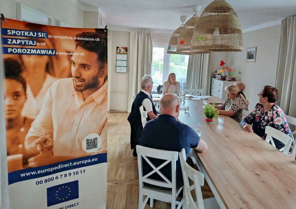 Wnętrze jasnego pomieszczenia, sześć osób siedzi przy długim drewnianym stole podczas spotkania. Po lewej stoi pionowy baner informacyjny, z tyłu duże okno i trzy wiszące plecione lampy.