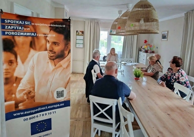 Wnętrze jasnego pomieszczenia, sześć osób siedzi przy długim drewnianym stole podczas spotkania. Po lewej stoi pionowy baner informacyjny, z tyłu duże okno i trzy wiszące plecione lampy.