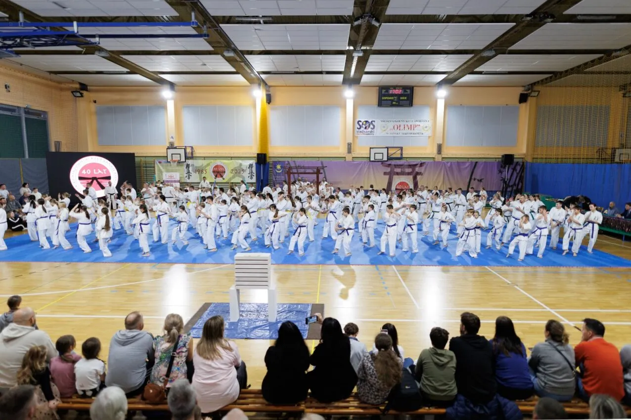 W sali gimnastycznej na niebieskiej macie ćwiczy około 60 osób w białych kimonach z niebieskimi pasami. Przed nimi na trybunach siedzi około 15 widzów oglądających pokaz. Na środku jest ustawiony stos białych desek do łamania. W tle wiszą banery z japońskimi motywami i informacją o 40-leciu klubu. Sufit jest podwieszany z widocznymi belkami świetlnymi.