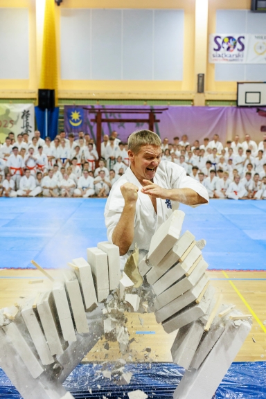 Mężczyzna w białym kimono uderza pięścią w stos białych cegieł łamiąc je na dwie części na hali sportowej, w tle widoczna grupa osób w białych strojach martial arts siedzących na niebieskiej macie.