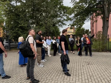 Grupa ludzi stoi na brukowanym placu, a mężczyzna w garniturze wskazuje w kierunku drzewa.