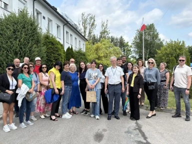 Grupa dorosłych osób stojących razem na chodniku przed budynkiem i drzewami.