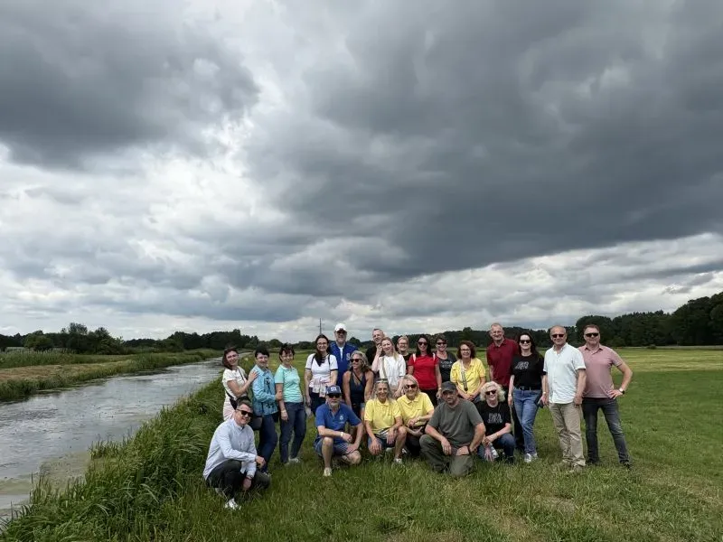 Grupa dorosłych osób pozująca na trawie nad rzeką pod zachmurzonym niebem.