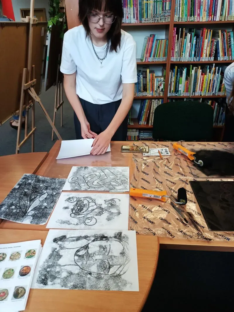 Kobieta w okularach stoi przy stole z czterema czarno-białymi rysunkami oraz narzędziami do malowania, na tle regału z książkami.