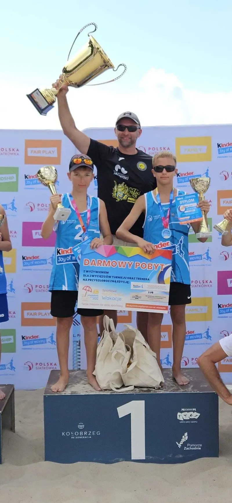 Dwóch chłopców i mężczyzna stoją na podium z numerem 1, trzymając puchary, dyplomy i duży plakat z napisem „DARMOWY POBYT”.