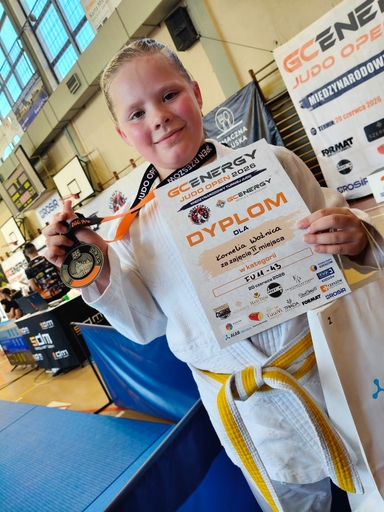Dziewczynka w białym kimonie z żółtym pasem stoi w hali sportowej. Trzyma medal na wstążce w lewej ręce i dyplom w prawej. Uśmiecha się przed matą i banerami turnieju.