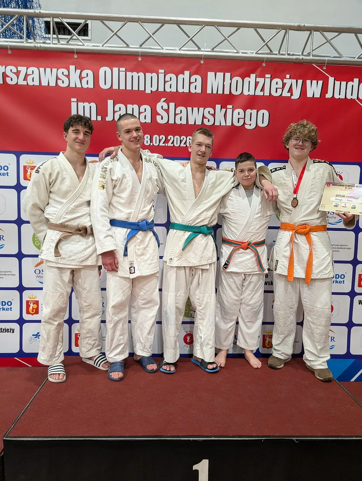 Pięciu młodych zawodników stoi razem na podium w białych judogach z różnokolorowymi pasami. Mają ręce na ramionach, jeden trzyma medal i dyplom; w tle duży czerwony baner i jasne wnętrze hali.