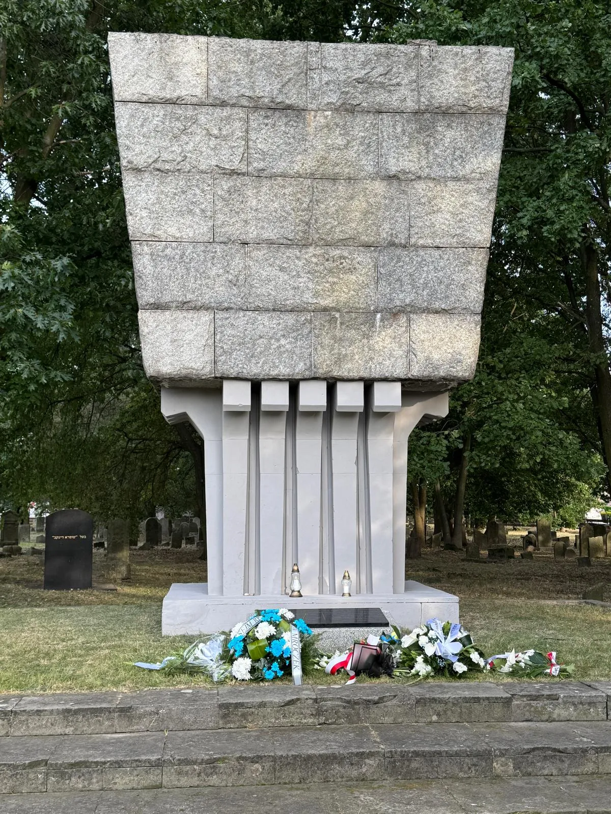 Pomnik zbudowany z jasnego kamienia z dekoracyjnymi pionowymi żłobieniami, przed którym leżą wieńce i kwiaty, na zewnątrz na tle drzew i macew.