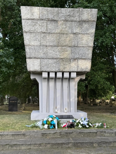 Pomnik zbudowany z jasnego kamienia z dekoracyjnymi pionowymi żłobieniami, przed którym leżą wieńce i kwiaty, na zewnątrz na tle drzew i macew.