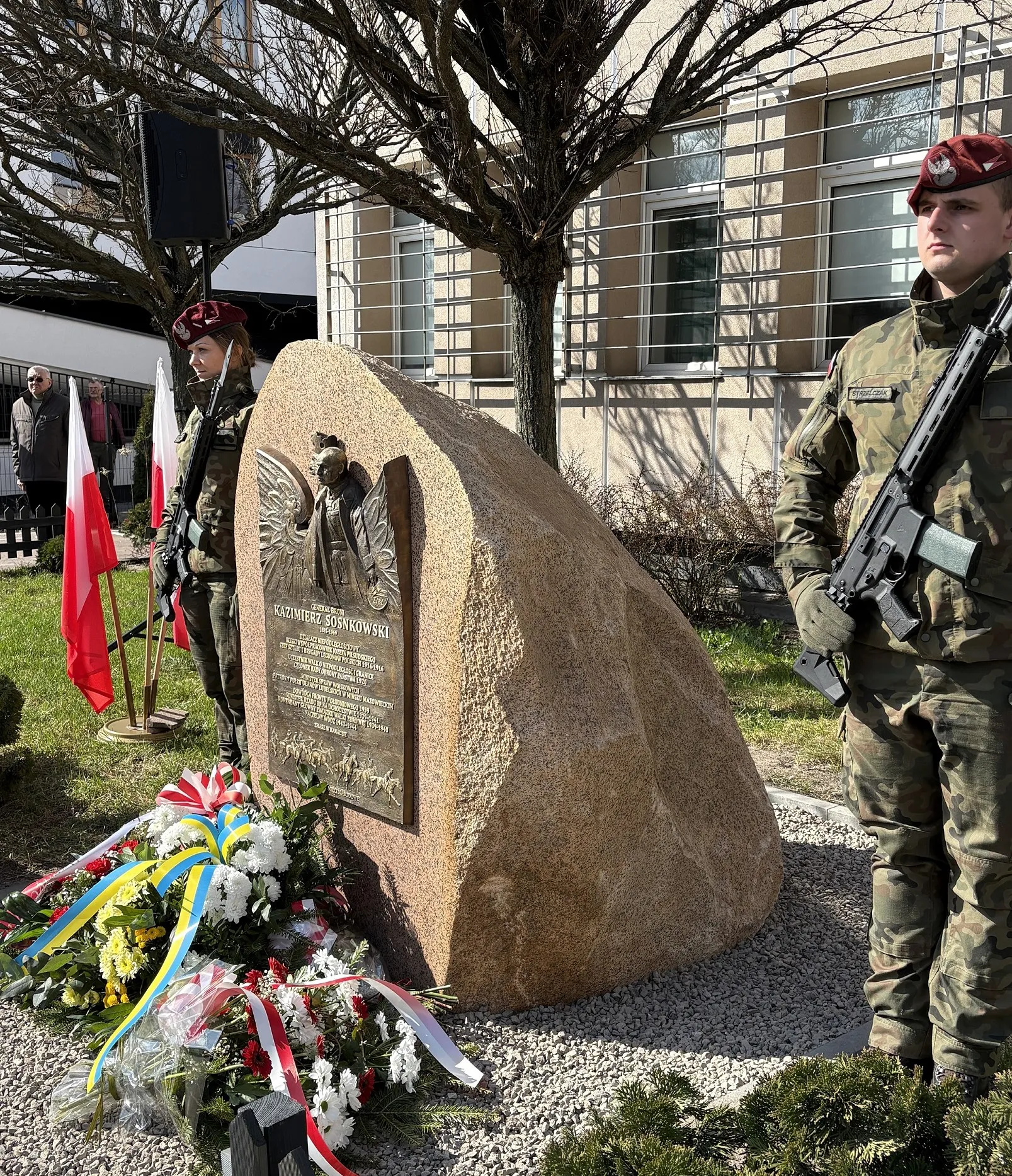 Duży kamienny pomnik z metalową tablicą ustawiony na żwirowym cokole. Przed pomnikiem leżą wiązanki kwiatów z biało-czerwonymi i niebiesko-żółtymi wstążkami. Po bokach stoją dwaj żołnierze w mundurach i czerwonych beretach z bronią. W tle nagie drzewa, budynek z oknami i kilka stojących flag Polski. Dzień, teren zewnętrzny.