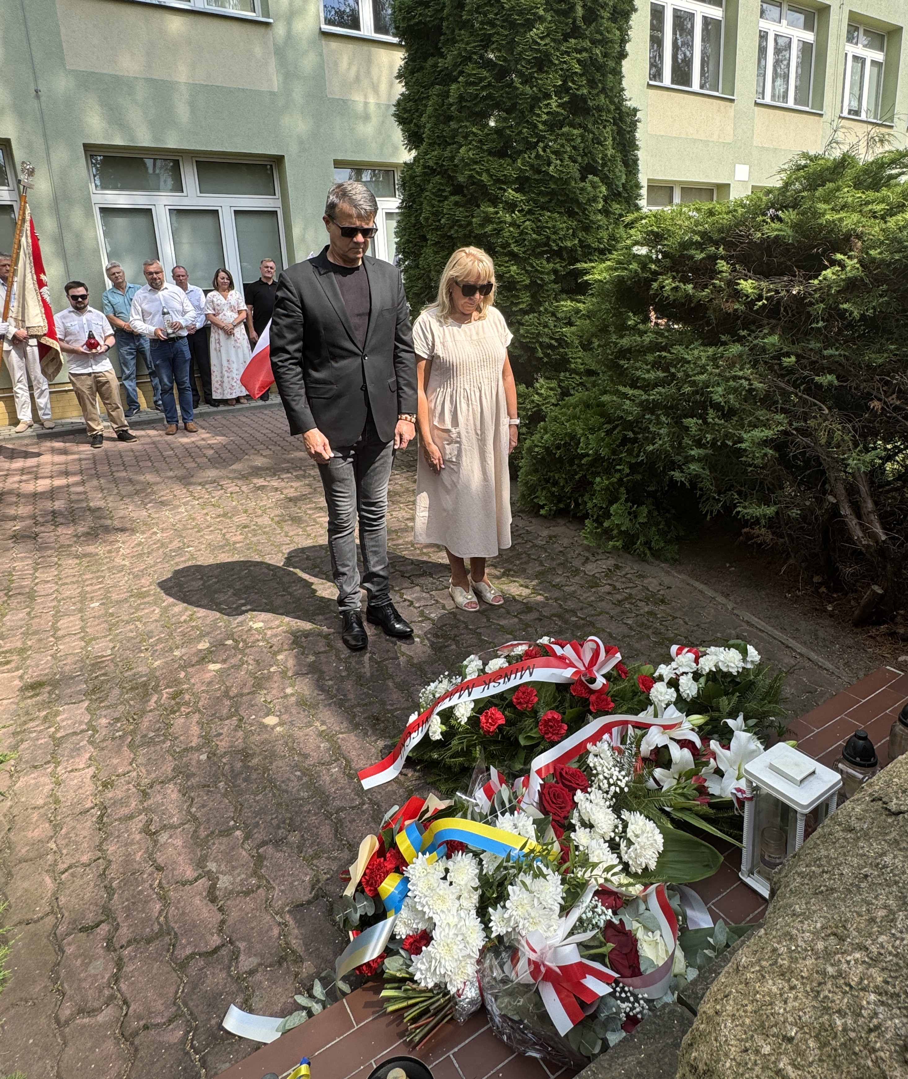 Mężczyzna w czarnym garniturze i kobieta w jasnej sukience stoją ze spuszczonymi głowami nad wiązankami kwiatów. Na pierwszym planie leżą czerwono-białe wieńce z wstążkami. W tle grupa ludzi przed zielonym budynkiem i wysokie krzewy.