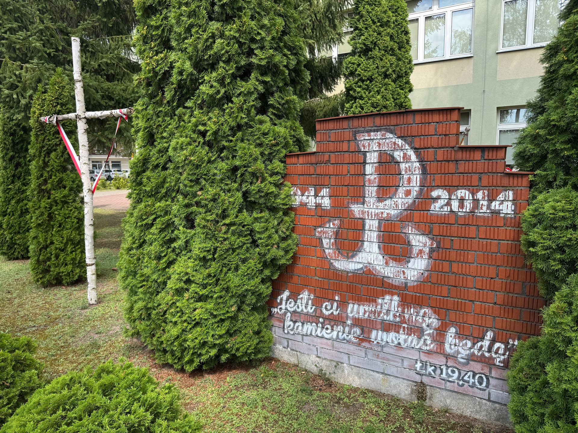 Biały krzyż z brzozowych pni stoi po lewej, owinięty biało-czerwoną wstążką. W centrum i po prawej wysokie zielone żywotniki zasłaniają część sceny. Po prawej czerwony mur z cegieł ma biały, częściowo starte malowidło, za nim widać jasnozielony budynek z oknami.