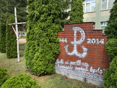 Biały krzyż z brzozowych pni stoi po lewej, owinięty biało-czerwoną wstążką. W centrum i po prawej wysokie zielone żywotniki zasłaniają część sceny. Po prawej czerwony mur z cegieł ma biały, częściowo starte malowidło, za nim widać jasnozielony budynek z oknami.
