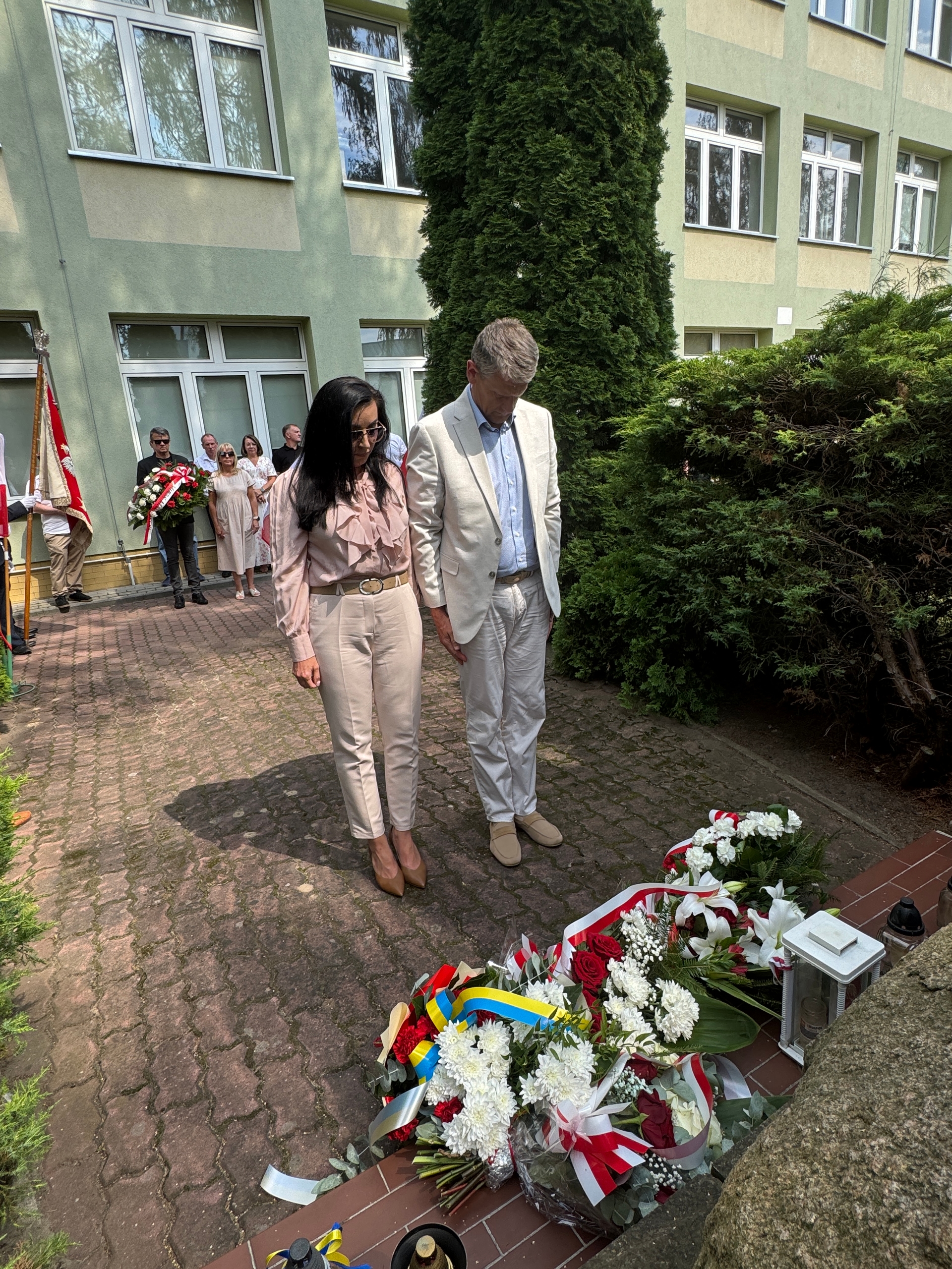 Dwoje dorosłych stoi przyklękniętych lub pochylonych z opuszczonymi głowami przed wieńcami kwiatów. W tle grupa osób przy budynku trzyma wieńce i sztandar. Otoczenie to dziedziniec przed zielonym budynkiem z drzewami i krzewami.
