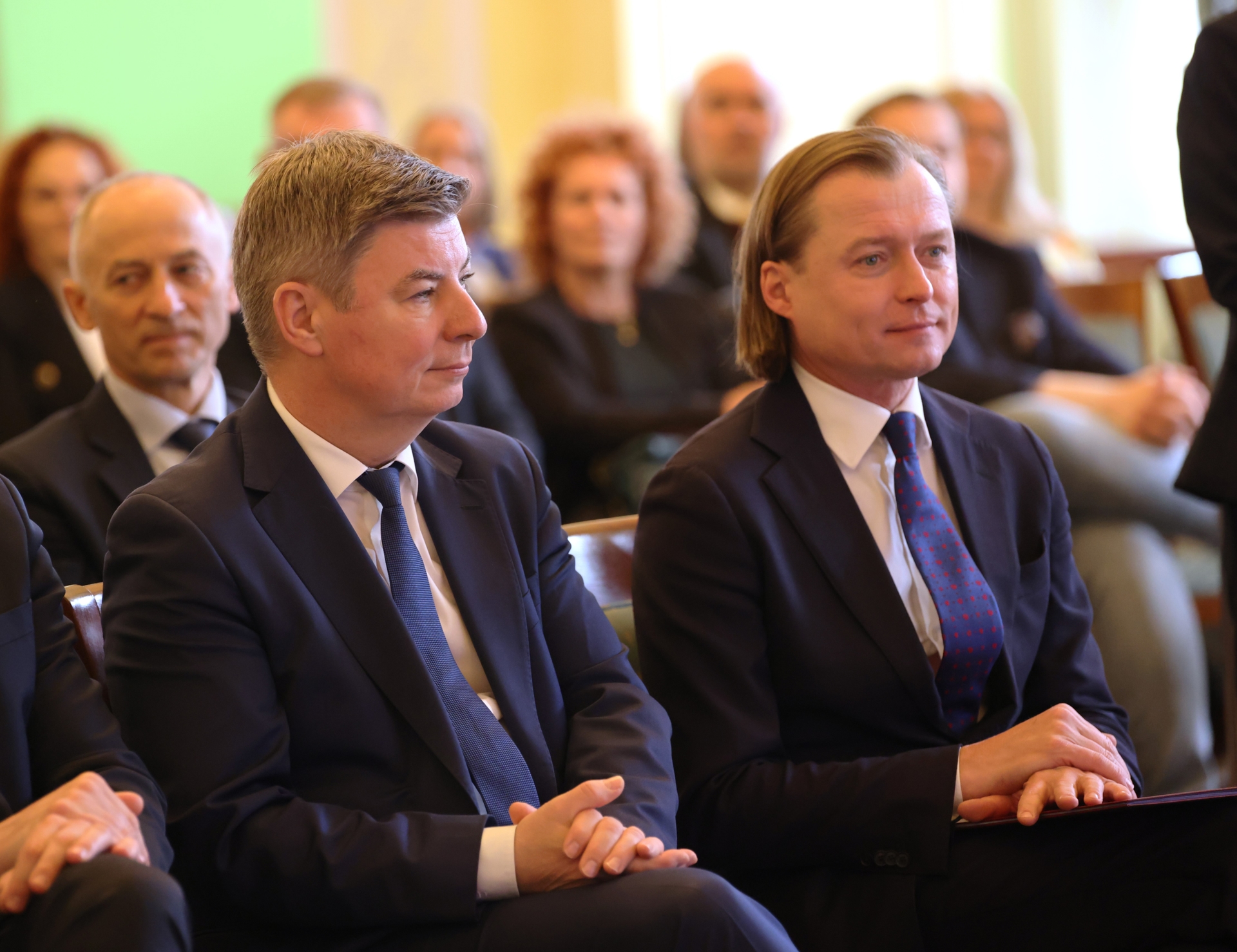 Dwóch mężczyzn w ciemnych garniturach siedzi w pierwszym rzędzie sali. W tle rozmyta grupa osób, jasne wnętrze i zielona ściana po lewej.
