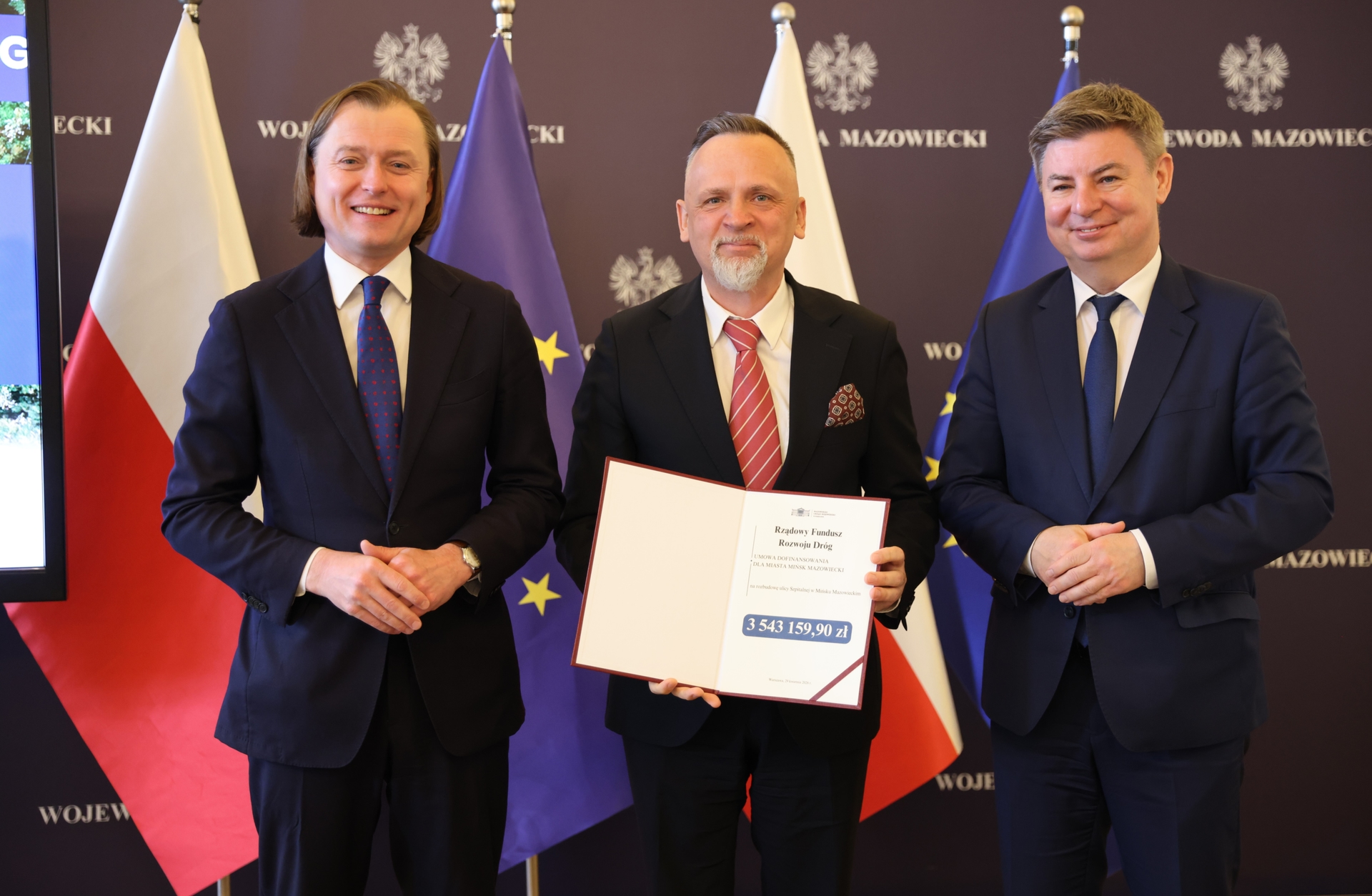 Trzech mężczyzn w garniturach stoi obok siebie wewnątrz budynku. W tle stoją flagi Polski i Unii Europejskiej na ciemnofioletowym tle z białymi godłami. Mężczyzna pośrodku trzyma otwarty dokument z widoczną sekcją dotyczącą kwoty.