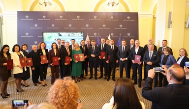 Grupa około 24 osób w formalnych strojach stoi w pomieszczeniu z żółtymi ścianami i kryształowymi żyrandolami. Osoby trzymają czerwone teczki, stoją przed ciemnobordowym panelem z herbami i flagami, a z przodu widać publiczność robiącą zdjęcia.