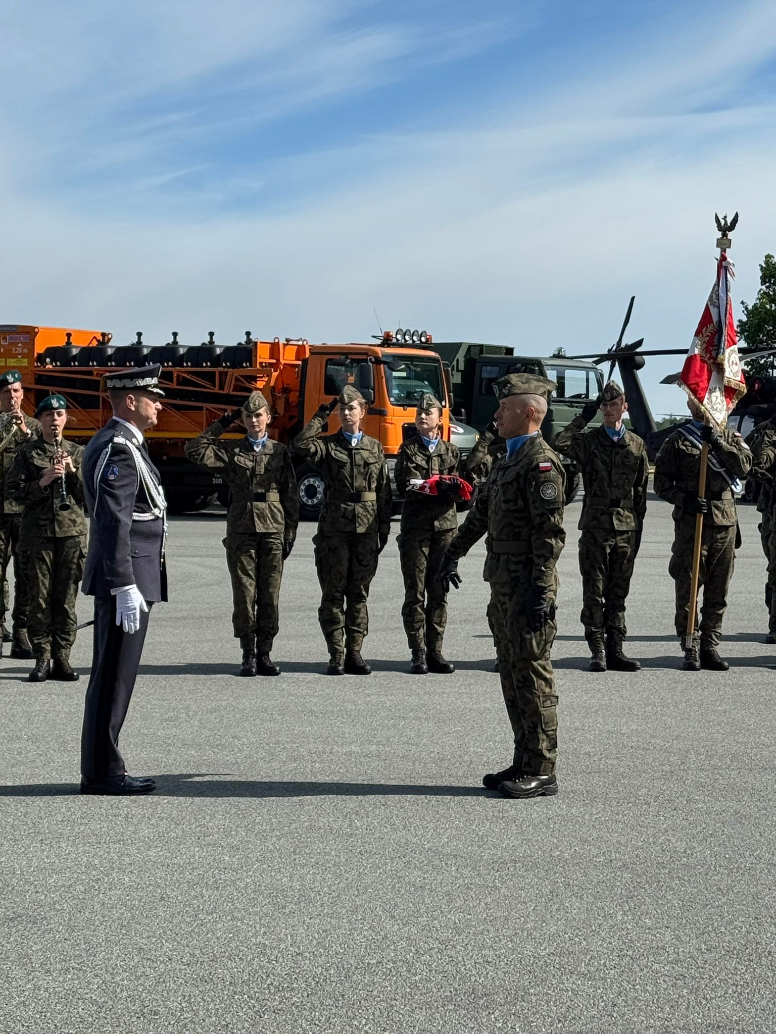 Grupa umundurowanych żołnierzy stoi na płycie lotniska i salutuje. Oficer w ciemnym mundurze stoi przodem do grupy. Z tyłu widać pomarańczowy samochód służbowy, wirnik helikoptera i biało-niebieskie niebo.