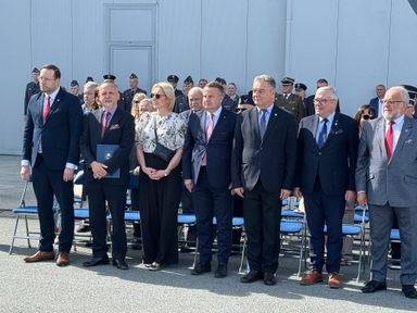 Siedmiu mężczyzn i jedna kobieta stoją w jednym rzędzie w garniturach, kobieta ma jasną bluzkę i okulary przeciwsłoneczne. Osoby stoją przed wysoką jasnoszarą ścianą budynku na zewnątrz w ciągu dnia. Za nimi stoi grupa umundurowanych oficerów, a przed nimi ustawione są niebieskie składane krzesła.