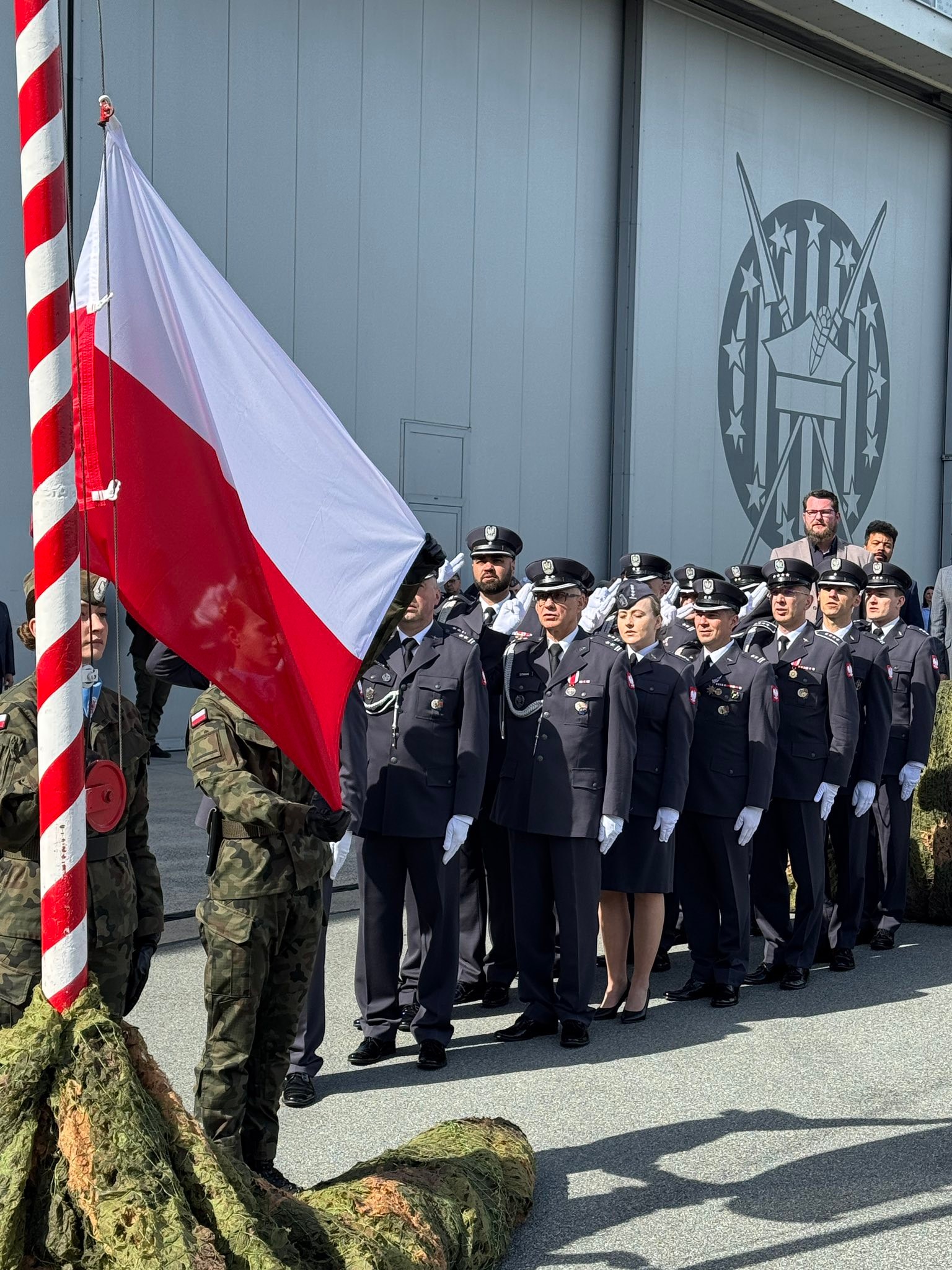 Polska flaga na maszcie uniesiona obok czerwono-białego słupa. Grupka umundurowanych funkcjonariuszy stoi w szeregu i oddaje salut przed hangarem z dużym emblematem na ścianie. Dwaj żołnierze w mundurach kamuflażowych trzymają flagę u dołu.