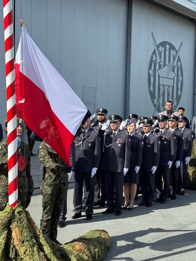 Polska flaga na maszcie uniesiona obok czerwono-białego słupa. Grupka umundurowanych funkcjonariuszy stoi w szeregu i oddaje salut przed hangarem z dużym emblematem na ścianie. Dwaj żołnierze w mundurach kamuflażowych trzymają flagę u dołu.
