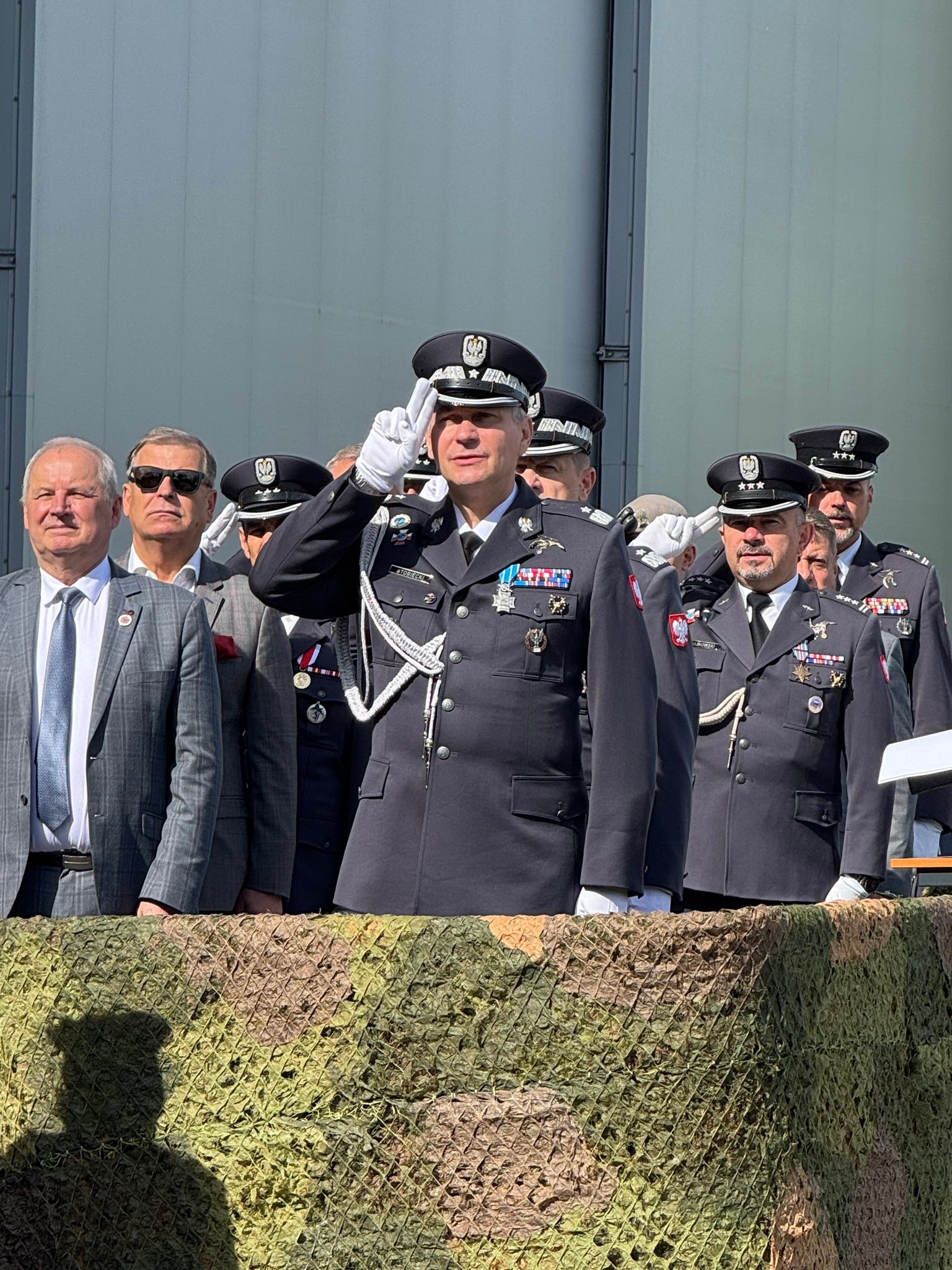 Grupa mundurowych i kilku mężczyzn w garniturach stoi na zewnątrz przed dużą szarą ścianą. Centralny oficer w ciemnym mundurze i białych rękawiczkach stoi z dłonią przy czole w salucie; na pierwszym planie zielono-brązowa siatka maskująca i cień postaci.