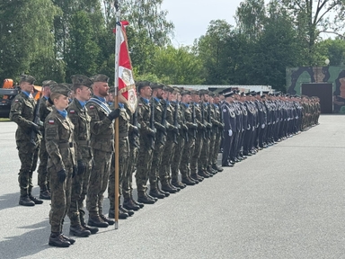 Żołnierze stoją w długim, równym szyku na zewnątrz w ciągu dnia. Po lewej żołnierz trzyma czerwono-biały sztandar. Większość ma mundury polowe z niebieskimi chustami, część ma ciemne mundury galowe, w tle drzewa i kontenery.