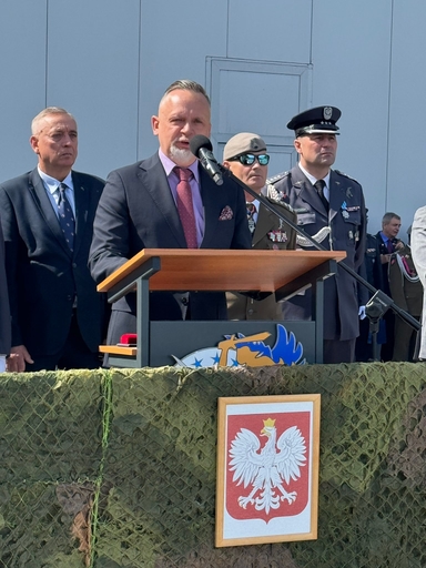 Mężczyzna w ciemnym garniturze i czerwonym krawacie przemawia przy mównicy do mikrofonu. Za nim stoją trzej mężczyźni w mundurach i garniturach. Przed mównicą wisi płótno maskujące, na którym opiera się oprawione godło Polski.