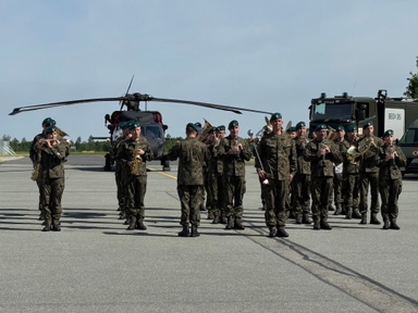 Grupa żołnierzy w mundurach i zielonych beretach stoi w szyku na lotniskowym płycie i gra na instrumentach dętych. Za nimi stoi śmigłowiec i wojskowe pojazdy na tle jasnego nieba.