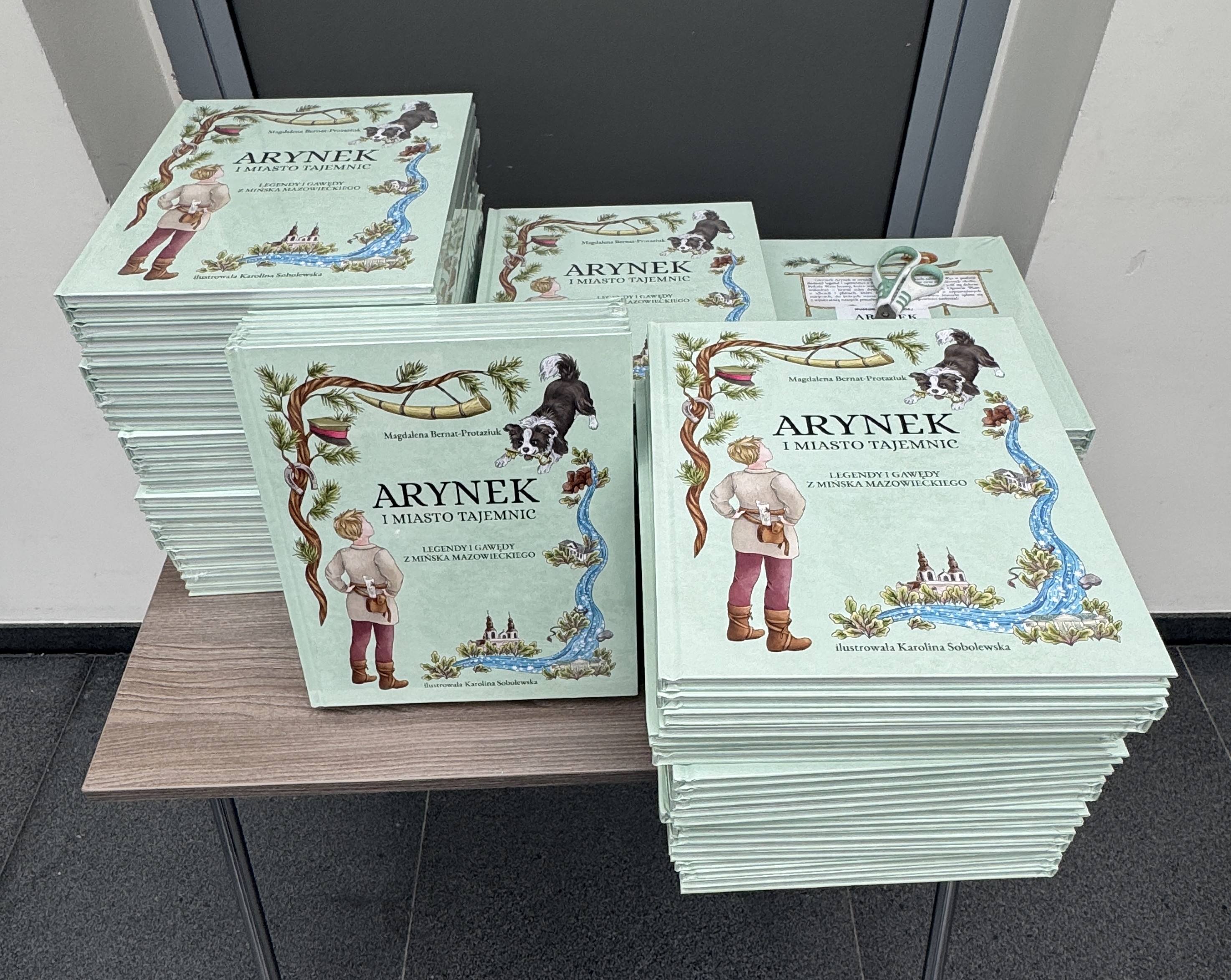 Stosy zielonkawych książek ułożone na drewnianym stole w pomieszczeniu. Kilka grubszych stosów obok siebie. Okładki z ilustracjami postaci stojącej tyłem, psem i motywem rzeki. Na jednym stosie leżą nożyczki. Tło ciemne drzwi i jasne ściany.