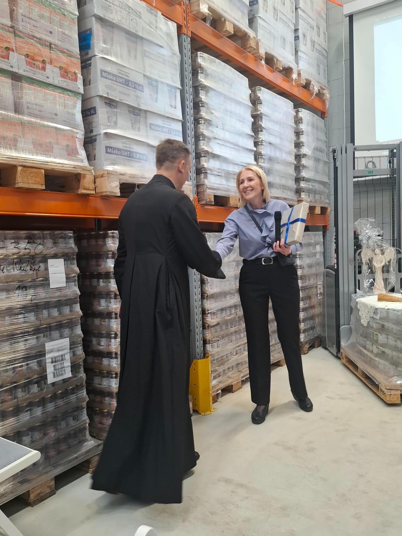 Kobieta trzyma mikrofon i opakowany prezent, podaje rękę mężczyźnie w czarnym stroju. Oboje stoją wewnątrz magazynu między wysokimi paletami owiniętymi folią.