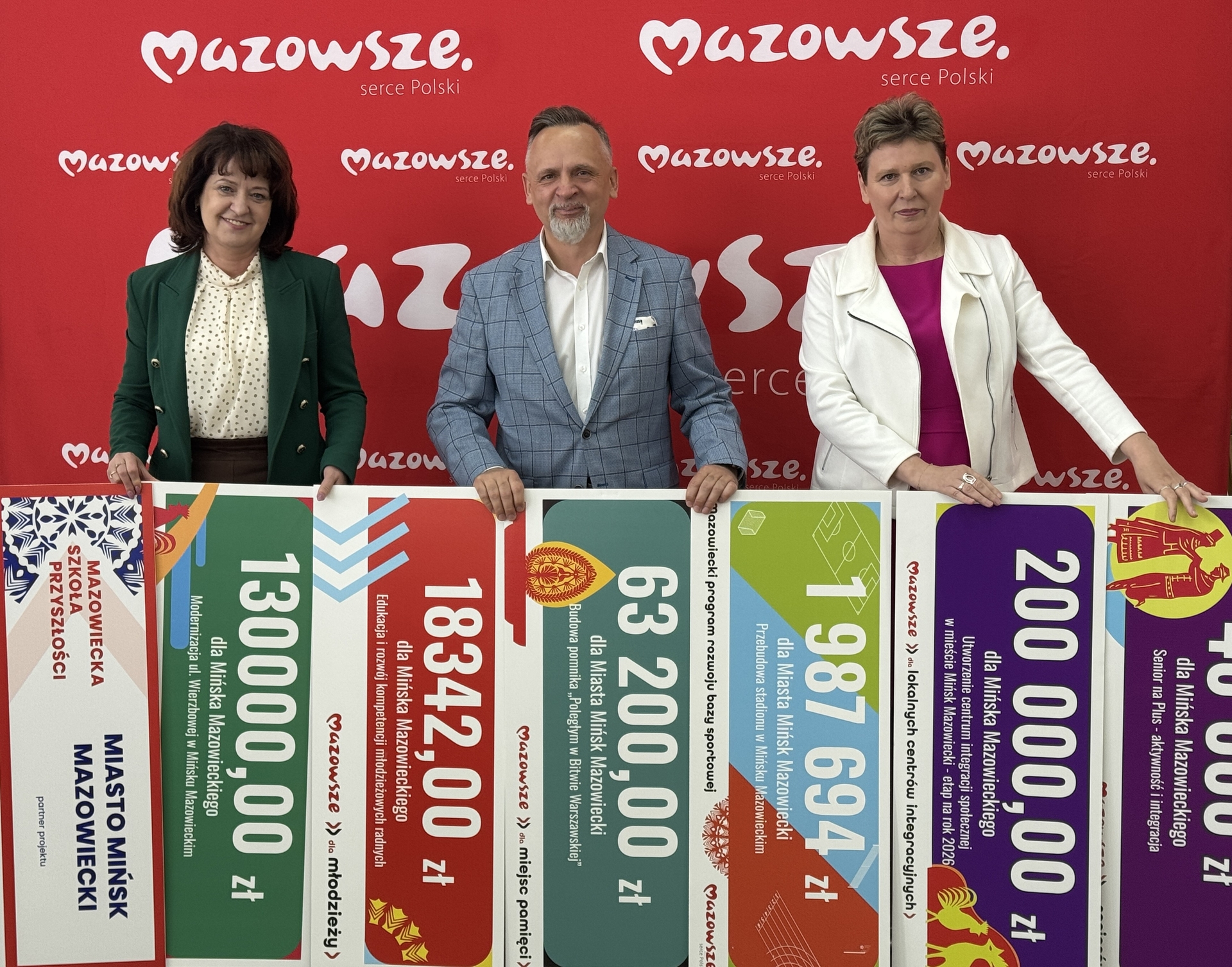 Trzy osoby stoją na czerwonym tle z białymi logotypami regionu. Trzymają poziome, kolorowe banery z dużymi liczbami i ikonami.