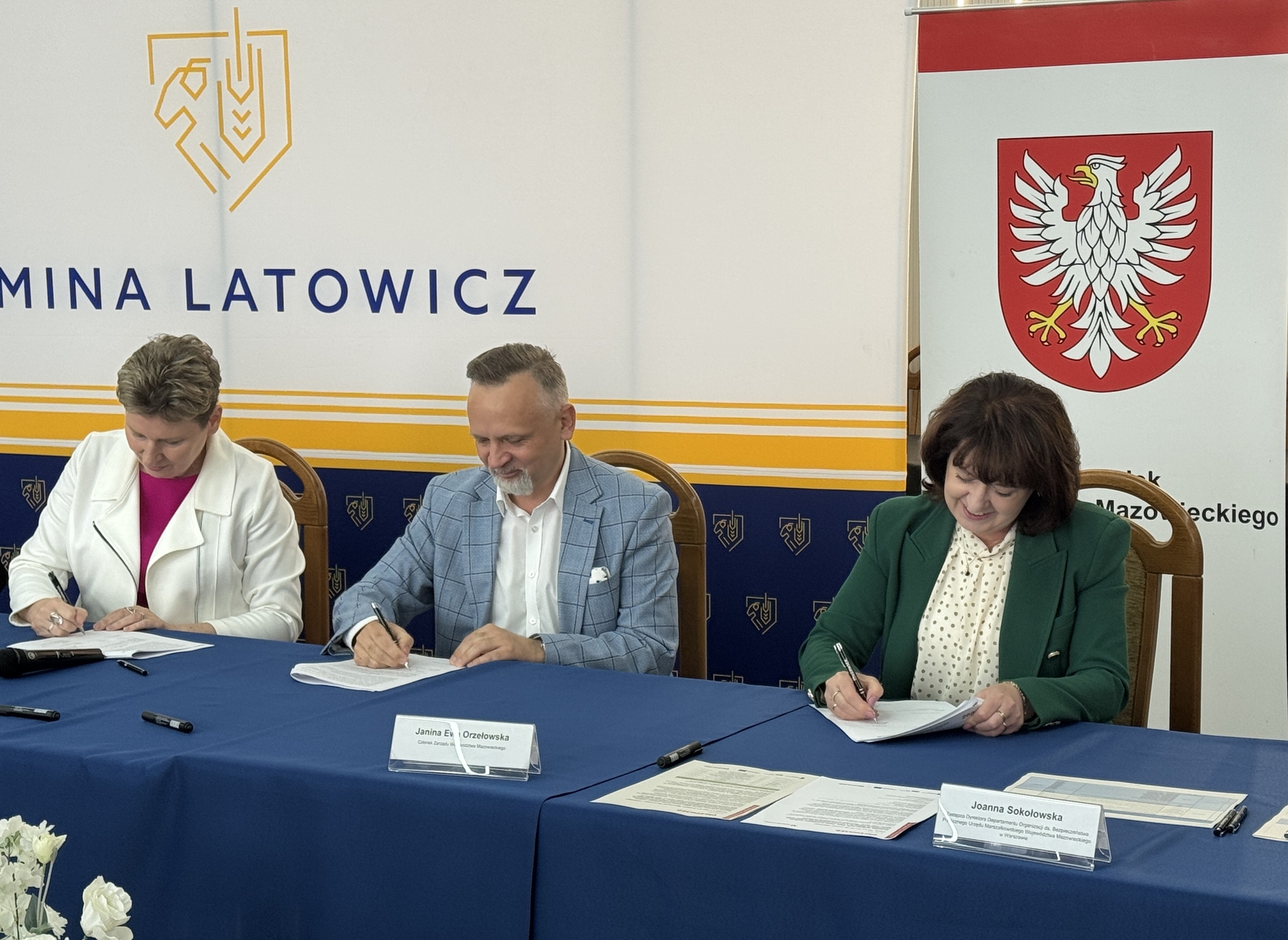 Trzy osoby siedzą przy niebieskim stole i podpisują dokumenty. W tle biało-żółto-granatowy baner z graficznym logotypem po lewej i czerwonym herbem z białym orłem po prawej. Wnętrze sali, widoczne krzesła i dokumenty na stole.